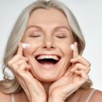 older woman smiling and spreading collagen cream on her cheeks